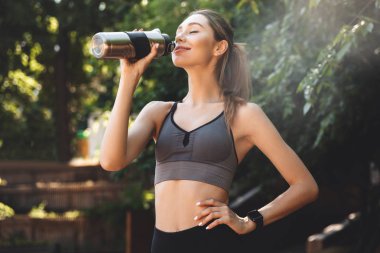 Gülümseyen genç fitness kız içme suyu bir şişe parkta duran