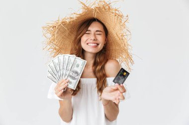 Beyaz arka plan üzerinde beyaz esmer kadın 20'li yaşlarda giyen büyük hasır şapka kredi kartı ve fan doları nakit tutarken gülümseyen fotoğrafını izole