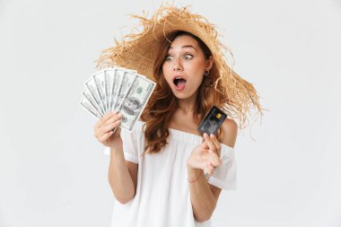 Heyecanlı güzel kadın 20'li yaşlarda giyen büyük hasır şapka kredi kartı ve beyaz arka plan üzerinde izole doları banknot hayranı tutarak fotoğrafı