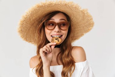 Görüntü closeup neşeli kadın 20'li yaşlarda giyen büyük hasır şapka ve güneş gözlüğü beyaz arka plan üzerinde izole bisküvi yemek