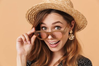 Neşeli genç kadın 20'li yaşlarda giyen hasır şapka ve güneş gözlüğü bej renkli arka plan üzerinde izole kamera bakarak portre