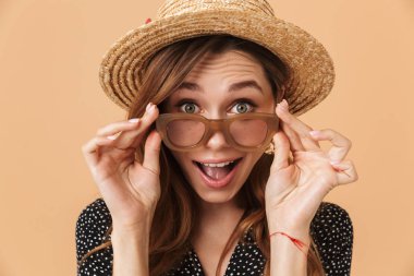 Güneş gözlüğü dokunmaktan ve bej renkli arka plan üzerinde izole kamera bakarak yaz sürpriz kadın 20'li yaşlarda giyen hasır şapka portresi