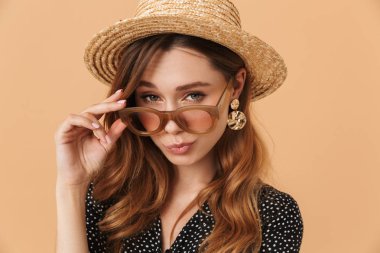 Modern moda kadın 20'li yaşlarda giyen hasır şapka ve güneş gözlüğü bej renkli arka plan üzerinde izole kamera bakarak portre