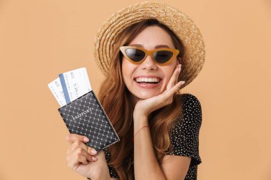 Şirin esmer kadın 20'li yaşlarda giyen hasır şapka ve güneş gözlüğü bej renkli arka plan üzerinde izole seyahat biletleri ile pasaport tutarken sevindirici