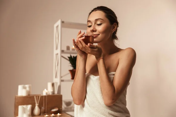 stock image Portrait of a delighted young woman wrapped in a towel drinking herbal tea in a glass at the spa center