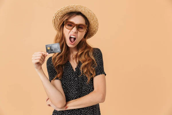 麦わら帽子と笑いと ベージュの背景の上分離されたクレジット カードを保持しているサングラスを着て満足している若い女性の画像 — ストック写真