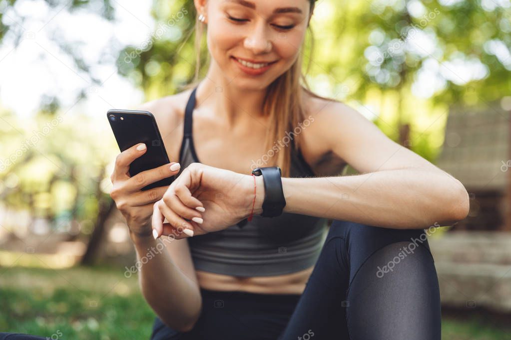 Cropped image of a smiling young fitness girl holding mobile phone while sitting outdoors and looking at smart watch
