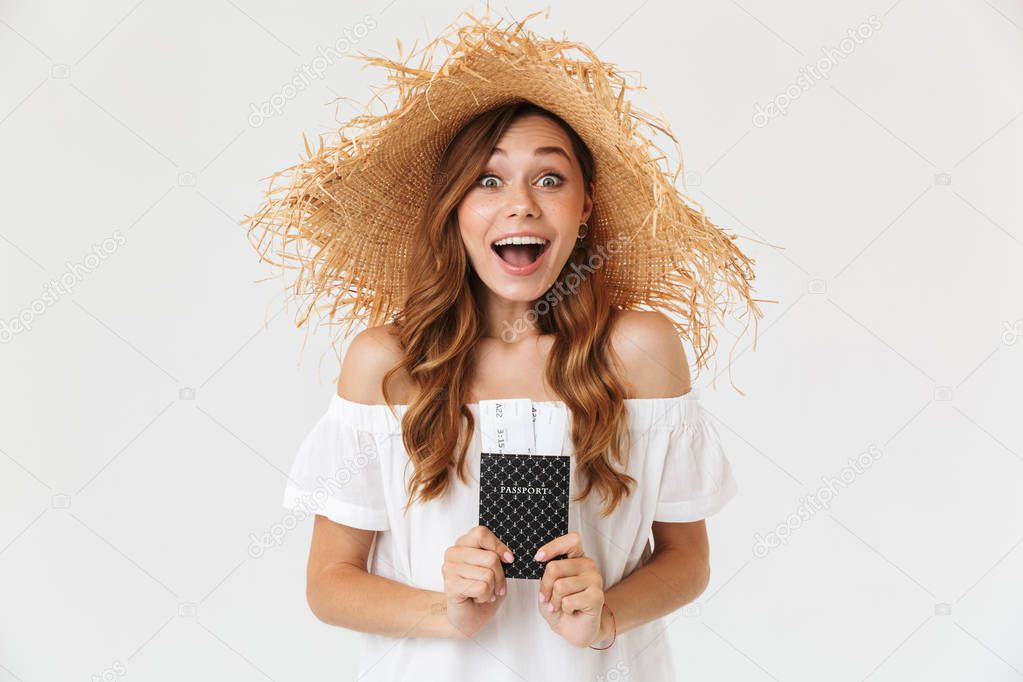 Portrait of excited summer woman 20s wearing big straw hat rejoicing while holding passport with tickets isolated over white background
