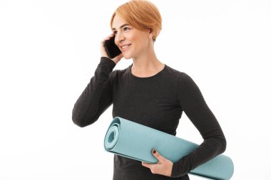 Gülümseyen bir yoga kadın portresi fitness mat beyaz arka plan üzerinde tutarak cep telefonu üzerinde konuşurken spor elbise giymiş