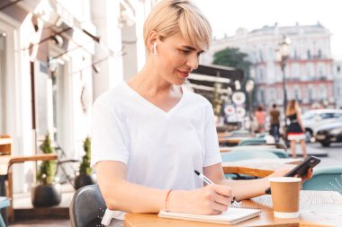 Hücre telefon ve kablosuz kulaklık kullanarak not defterinde yazarken açık yaz kafede oturan beyaz t-shirt giyen beyaz ciddi kadın fotoğrafı