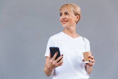 Kağıt bardak ve gri duvardan izole smartphone kahve tutarken casual giyim gülümseyen giyen neşeli sarışın kadın görüntüsünü