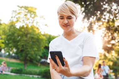 Yeşil şehir parkta yürüyüş sırasında smartphone tutarken casual giyim gülümseyen giyen neşeli sarışın kadın görüntüsünü