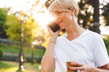 Süre smartphone üzerinde konuşmak ve yeşil parkta yürüyüş sırasında paket servisi olan restoran kahve tutan casual giyim gülümseyen giyen memnun çekici kadın görüntüsünü