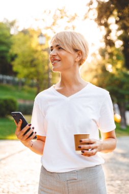 Yeşil Park yürüyüş sırasında smartphone ve paket servisi olan restoran kahve tutarken casual giyim gülümseyen giyen neşeli sarışın kadın görüntüsünü