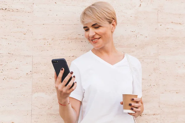 照片特写的快乐金发女人穿着白色 T恤衫使用手机 而站在反对米色墙室外夏季和喝咖啡从纸杯 — 图库照片