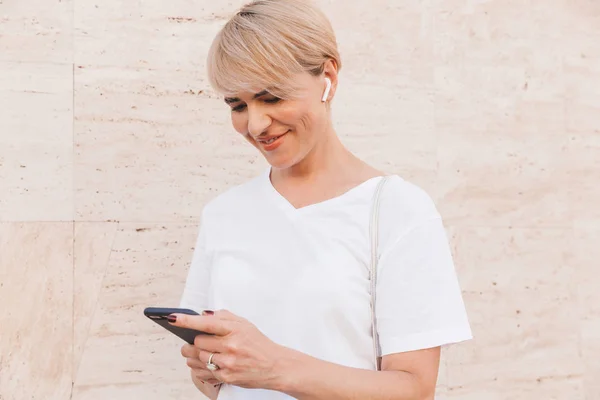 Photo of happy blond woman wearing white t-shirt using mobile phone while standing against beige wall outdoor with bluetooth earphone