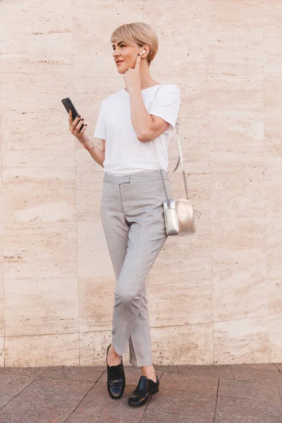 Full length image of stylish blond woman 30s wearing white t-shirt using mobile phone while standing against beige wall outdoor and touching bluetooth earphone