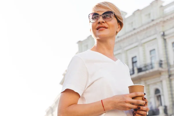 微笑的白种金发女人30S 穿着白色 T恤和太阳镜穿过城市街道与外卖咖啡 — 图库照片