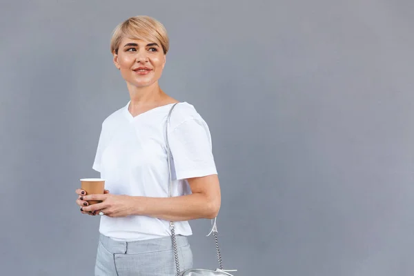 内容成熟妇女的照片穿着白色 T恤站在灰墙上 从纸杯里拿咖啡 — 图库照片