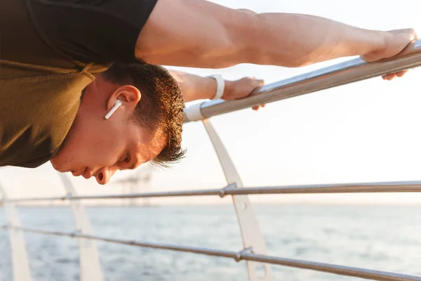 Concentrated young sportsman in earphones doing stretching exercises at the seaside, leaning on rails