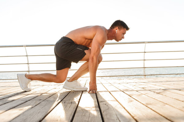 Focused young shirtless sportsman in earphones getting ready to run at the seaside