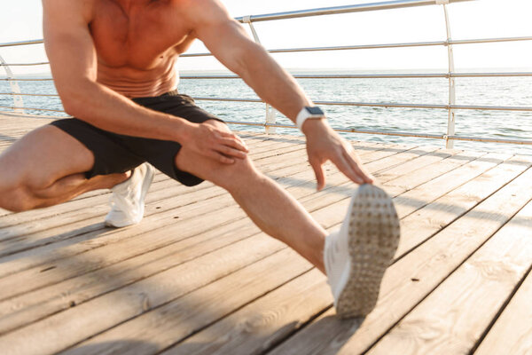 Cropped image of shirtless sportsman doing stretching leg exercises at the seaside