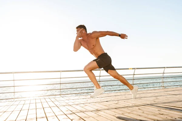 海辺で実行しているイヤホンの強い若い上半身裸スポーツマン — ストック写真