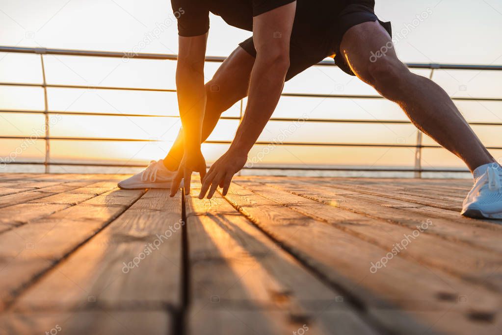 Cropped image young fit sportsman doing stretching leg exercises at the seaside at sunset