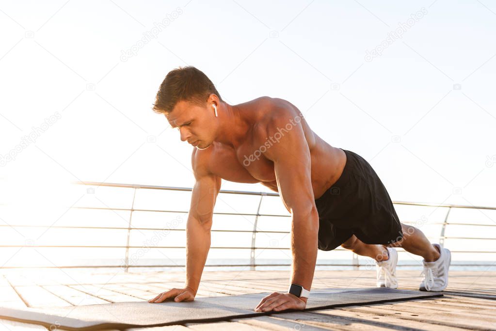 Image of handsome young sportsman outdoors on the beach make exercises on sport carpet listening music.