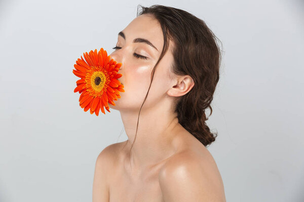 Beauty portrait of an attractive young topless woman with make-up posing with red gerbera flower at her face isolated over gray background