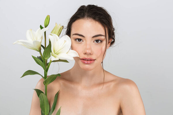 Beauty portrait of a lovely young topless woman with make-up posing with lily flower at her face isolated over gray background