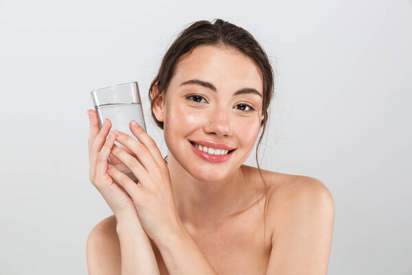 Beauty portrait of a smiling young topless woman with make-up holding glass of water isolated over gray background