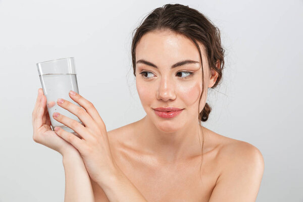 Beauty portrait of a lovely young topless woman with make-up holding glass of water isolated over gray background