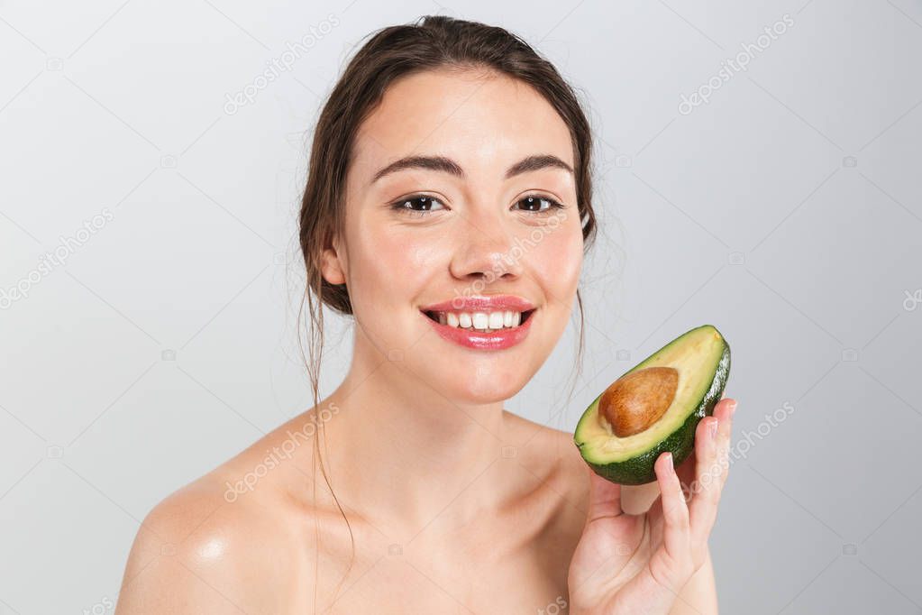 Beauty portrait of a cheerful young topless woman with make-up holding sliced avocado isolated over gray background