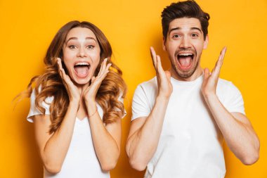 Photo of emotional couple man and woman in basic clothing screaming in surprise or delight and raising arms isolated over yellow background clipart