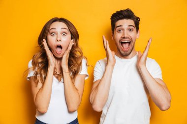 Photo of excited couple man and woman in basic clothing shouting in surprise or delight and raising arms isolated over yellow background clipart