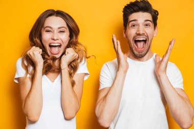 Photo of beautiful admired couple man and woman in basic clothing screaming in surprise or delight and touching cheeks isolated over yellow background clipart