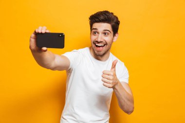 Başparmak gösterilen sarı arka plan üzerinde izole cep telefonunuzla bir sefie alarak neşeli genç bir adam portresi