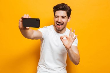 Tamam gösterilen sarı arka plan üzerinde izole cep telefonunuzla bir sefie alarak neşeli genç bir adam portresi