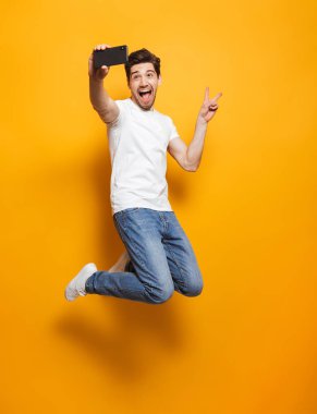 Tam uzunlukta atlama ve barış işareti izole sarı arka plan üzerinde siyah Smartphone cep telefonu ile selfie çekerken gösterilen kahverengi saçlı memnun adam portresi