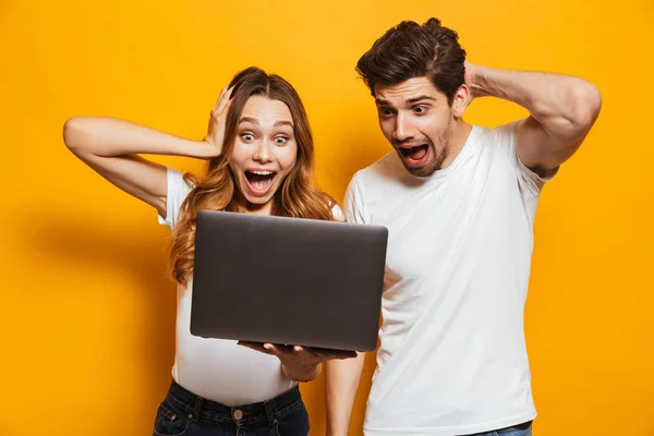 Portrait of excited couple man and woman screaming and grabbing head while holding black laptop isolated over yellow background