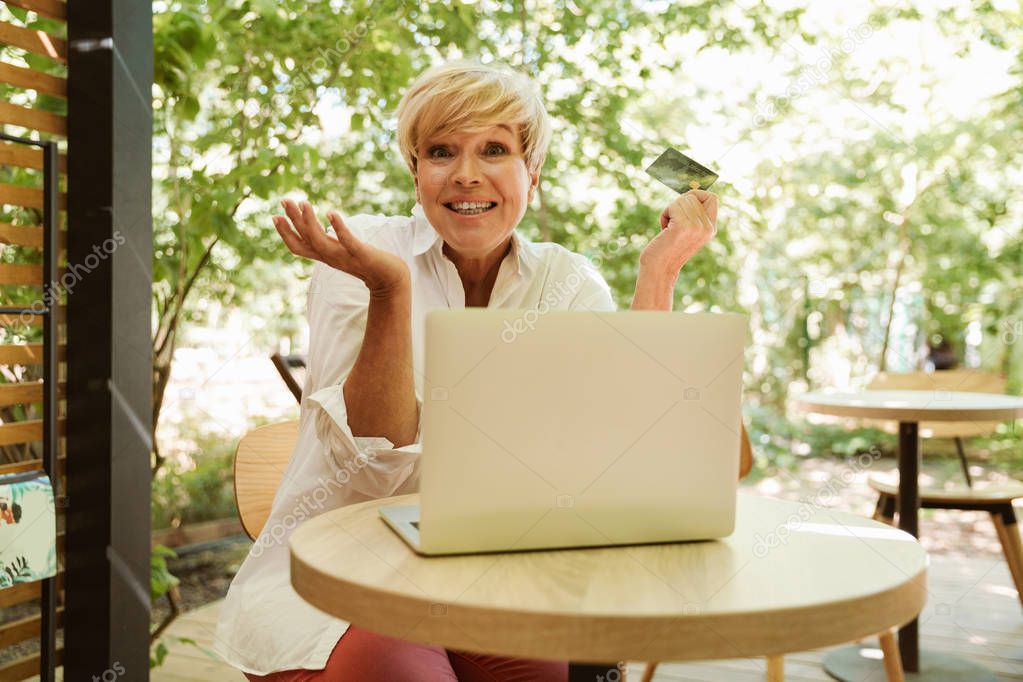 Excited mature woman sitting at a cafe with laptop computer and showing plastic credit card