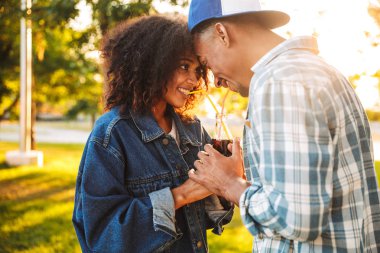 Gülümseyen genç Afrikalı çift birlikte parkta ayakta iken iki payet ile gazlı içecek ile bir şişeden içme portresi
