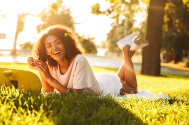 Mutlu genç Afrikalı kadın görüntüsünü çim açık havada kaykay parkı üzerinde yatıyor.