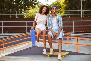 Genç, neşeli bir Afrikalı çift ile birlikte paten parkta oturan kaykaylar portresi