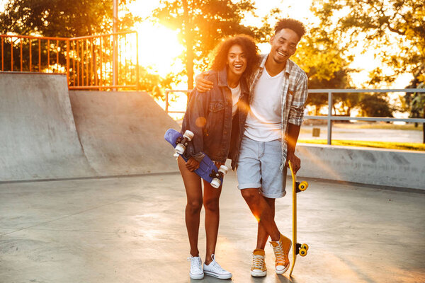 Portrait of a laughing young african couple with skateboards standing together at the skate park