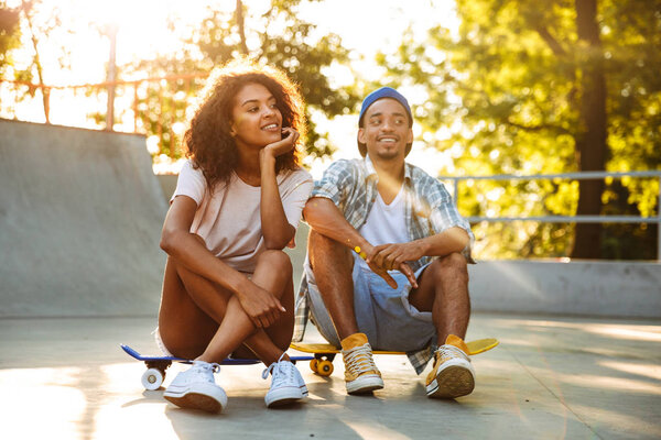 Portrait of a happy young african couple with skateboard sitting together at the skate park