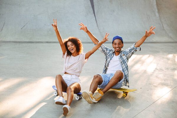 Portrait of a happy young african couple with skateboards sitting together at the skate park with hands outstretched