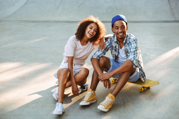 Portrait of a smiling young african couple with skateboards sitting together at the skate park