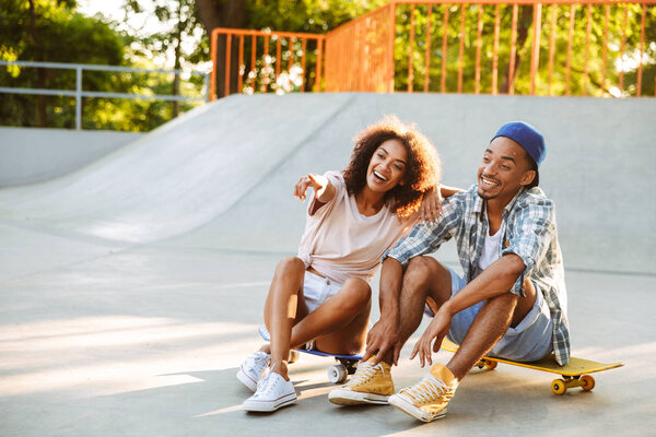 Portrait of a happy young african couple with skateboards sitting together at the skate park pointing finger